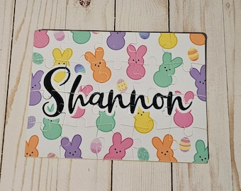 May include: A personalized jigsaw puzzle featuring the name "Shannon" in black script. The puzzle pieces are decorated with colorful Easter-themed graphics, including bunny-shaped marshmallow treats and decorated eggs. The background is white.