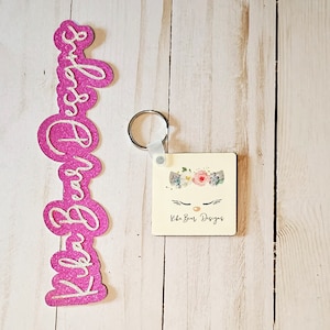 May include: A pink glittery Kika Bear Designs keychain with a square design featuring a unicorn face with a floral crown. The keychain is attached to a silver keyring. A matching pink glittery Kika Bear Designs cutout is also present.
