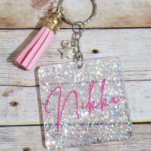 May include: A square keychain featuring a clear acrylic base filled with iridescent silver glitter. The name Nikki is printed in bright pink script across the center. A small silver star charm and a light pink faux leather tassel are attached.