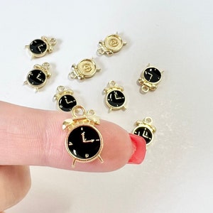 Puede incluir: Un grupo de diez dijes de reloj despertador de color dorado con carátulas de reloj negras. Los dijes tienen un bucle en la parte superior para sujetarlos a un collar o pulsera.
