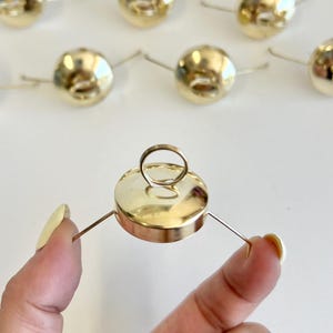 May include: A collection of small, shiny, gold-colored metal ornaments. Each ornament has a round, bulbous shape with a small loop at the top and thin wire extensions. One ornament is held in a hand, showcasing its design. The ornaments are likely for decoration.