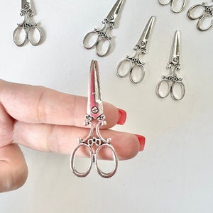May include: A set of ten miniature silver-toned scissors with ornate details. The scissors are arranged in a scattered pattern on a white surface. One scissor is being held by a hand.