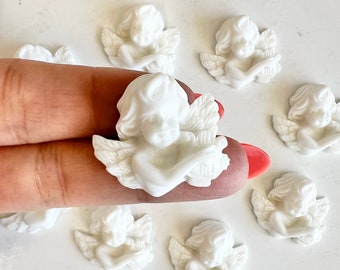May include: A collection of white cherub figurines, each with detailed wings and a serene expression. The cherubs are arranged on a white surface, with one held by a hand. These decorative figurines are ideal for crafts or home decor.