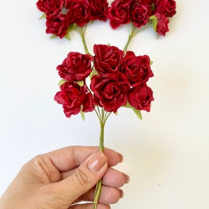 May include: A bouquet of small, red paper roses with green stems. The roses are arranged in a cluster and are held in a hand.