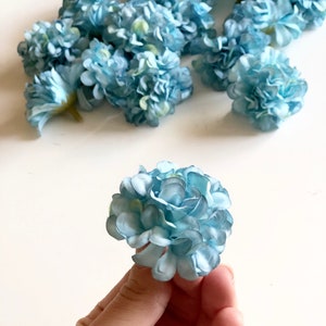 Op de afbeelding: Een tros lichtblauwe kunstbloemen, elke bloem is een kleine bol van bloemblaadjes. Eén bloem wordt in een hand gehouden.