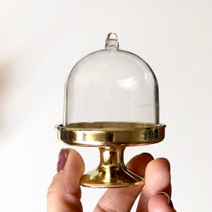 May include: A small, clear glass cake dome with a gold metal base. The dome is on a stand with a round base.