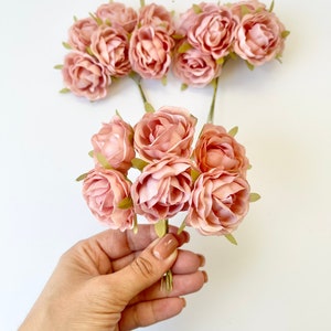 May include: A bunch of blush pink paper roses with green stems. The roses are arranged in a bouquet and are held in a hand.