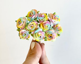 May include: A bunch of rainbow paper roses with green stems. The roses are arranged in a bouquet and are held in a hand.