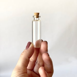 May include: A clear glass bottle with a cork stopper. The bottle is small and is being held in a hand.