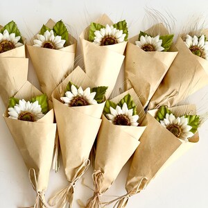 May include: A collection of ten small bouquets of white faux sunflowers with green leaves, wrapped in brown paper cones and tied with twine.