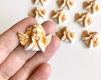 May include: A collection of small, white and gold angel figurines. The angels have their hands on their faces, with detailed wings and hair. The figurines are arranged on a white surface.