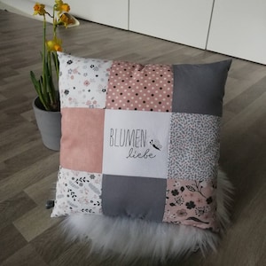 Op de afbeelding: Een grijs en roze patchwork kussen met de tekst "BLUMEN liebe" geborduurd op de voorkant. Het kussen heeft verschillende bloemmotieven.