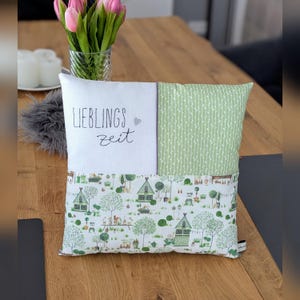 Könnte beinhalten: Ein dekoratives quadratisches Kissen mit Patchwork-Design. Der obere linke Bereich ist weiß mit dem Text "LIEBLINGS zeit" in schwarzer Schrift. Der obere rechte Bereich ist hellgrün mit einem vertikalen Streifenmuster. Der untere Bereich zeigt eine grün-weiße Dorflandschaft.