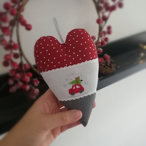 Op de afbeelding: Rood en wit hartvormig kussen met een wit geborduurd ontwerp van een rode auto die een groene kerstboom vervoert. Het kussen is versierd met witte stippen.