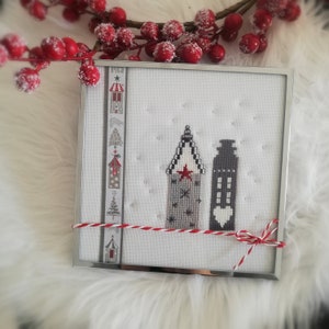 Op de afbeelding: Een ingelijste kruissteek met twee gestileerde gebouwen in grijstinten, zwart en wit, met een rode ster en hart. Het kunstwerk is geaccentueerd met een decoratief lint en een rood-witte touwstrik. De lijst is zilver.