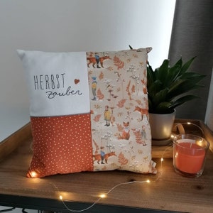 Op de afbeelding: Vierkant decoratief kussen met een patchwork ontwerp. Eén sectie heeft de geborduurde woorden "HERBST Zauber" met een hart. De andere secties hebben een roestkleurig stippenpatroon en een beige patroon met vossen, kinderen en bladeren.