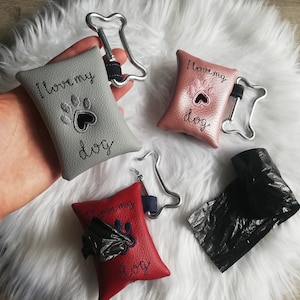 Może przedstawiać: Trzy uchwyty na worki na psie odchody z nadrukiem łapy i sercem. Uchwyty są wykonane ze skóry i dostępne są w kolorze szarym, różowym i czerwonym. Tekst na uchwytach brzmi "Kocham mojego psa".