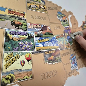 May include: A scratch-off map of the United States with illustrated state images. The map is tan with black outlines and state names. A hand is scratching off a state, revealing a colorful image. The map includes images of a bison, hot air balloons, and windmills.