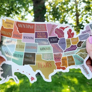 May include: A sticker of the United States of America with each state outlined and labeled with its name. The sticker is in the shape of the United States and is being held by two hands. The states are colored in different shades of pink, blue, green, yellow, orange, and brown.