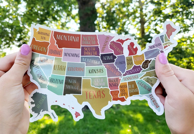May include: A sticker of the United States of America with each state outlined and labeled with its name. The sticker is in the shape of the United States and is being held by two hands. The states are colored in different shades of pink, blue, green, yellow, orange, and brown.