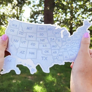 May include: A white sticker of the United States of America with each state outlined and labeled. The sticker is being held by two hands in front of a blurred green background.
