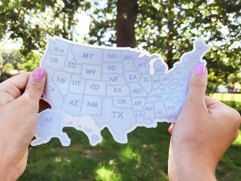 May include: A white sticker of the United States of America with each state outlined and labeled. The sticker is being held by two hands in front of a blurred green background.