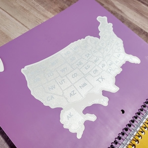 May include: A silver glitter sticker of the United States of America with each state labeled by its abbreviation. The sticker is on a purple notebook with a yellow spiral binding.
