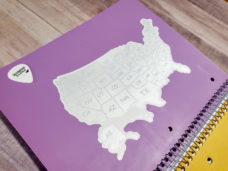 May include: A silver glitter sticker of the United States of America with each state labeled by its abbreviation. The sticker is on a purple notebook with a yellow spiral binding.