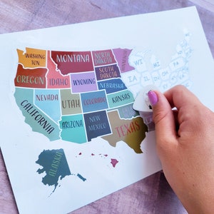 May include: A white map of the United States with each state outlined in a different color. The states are labeled with their names. A hand is holding a guitar pick over the state of Iowa.