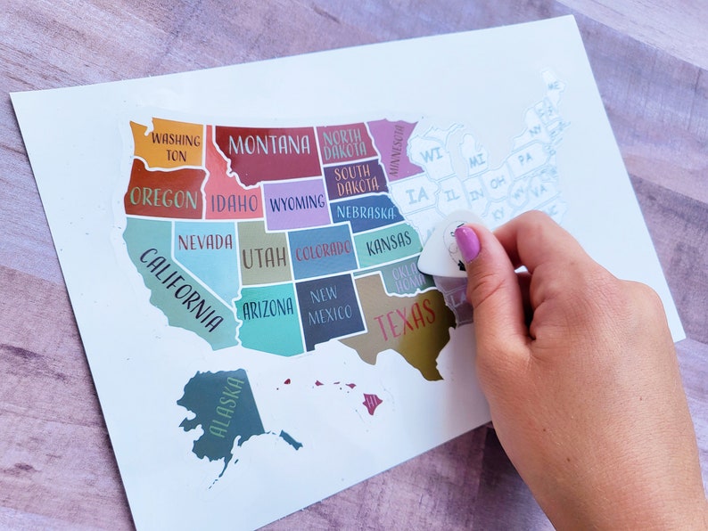 May include: A white map of the United States with each state outlined in a different color. The states are labeled with their names. A hand is holding a guitar pick over the state of Iowa.