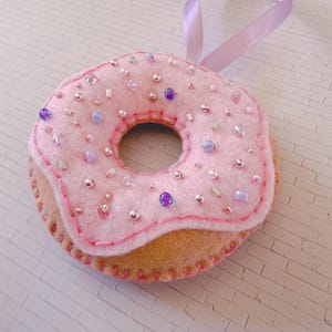 May include: A handmade felt donut decoration with a pink iced top and a tan base. The pink icing is decorated with small silver and purple beads. A lilac ribbon is attached for hanging.