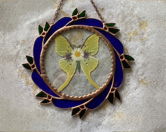 Puede incluir: Un atrapasol de vitral con un diseño de corona azul y verde. Una mariposa amarilla con una flor blanca está dentro de la corona.