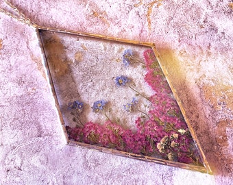 以下が含まれることがあります： 金色のダイヤモンド型のペンダントネックレス。中には紫と青の花が押し花で飾られています。ペンダントは、ざらざらしたピンクの表面の上に置かれています。