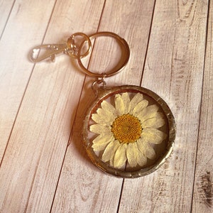 May include: A gold-toned keychain with a round, clear resin charm featuring a pressed white daisy with a yellow center.