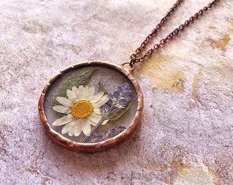 May include: A copper-toned pendant necklace featuring a clear, round glass pendant. Inside the pendant are a white daisy with a yellow center, lavender, and green leaves. The chain is also copper-toned.