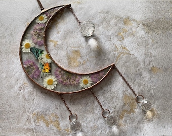 Puede incluir: Un adorno de pared en forma de media luna con un marco color cobre. La luna está llena de flores secas, margaritas y una mariposa. Cuentas de cristal transparente cuelgan de la parte inferior y una más grande de la parte superior.