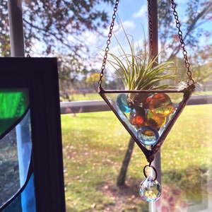 May include: A triangular glass terrarium with a copper chain hanging from a window. The terrarium contains a small air plant and colorful glass marbles. A crystal prism hangs from the bottom of the terrarium.