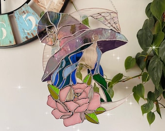 May include: A stained glass window hanging with a crescent moon design. The moon is decorated with a witch wearing a purple hat and a pink rose.