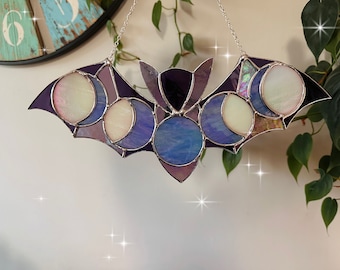 May include: A stained glass bat with a purple and blue design featuring a crescent moon pattern. The bat is hanging from a silver chain.
