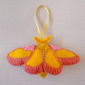 Könnte beinhalten: Handgefertigter Filz-Motten-Ornament mit gelben, rosa und goldenen Akzenten. Die Motte hat gelbe Flügel mit rosa und orangefarbenen Verzierungen, rosa Pailletten-Details und einen goldperlenbesetzten Körper. Ein gelbes Band ist zum Aufhängen befestigt.