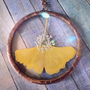 May include: A yellow ginkgo leaf and white flowers are encased in a round copper frame, creating a delicate and natural pendant.