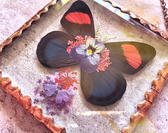 May include: A copper framed pendant with a black and red butterfly and pressed flowers inside. The butterfly has red accents on its wings and the flowers are purple, pink, and white.