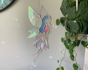May include: A stained glass fairy with iridescent wings and a pink dress. The fairy is holding a flower in her hand and is looking down.