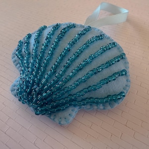 May include: A light blue felt seashell ornament with turquoise beaded accents. The beaded lines radiate from the center, mimicking the natural lines of a seashell. A white ribbon is attached for hanging.
