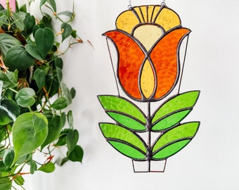 May include: A stained glass window hanging features a stylised orange and yellow flower with green leaves.
