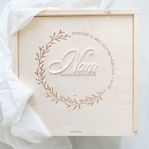 May include: A light-coloured wooden box with a lid. The lid features a decorative wreath design with the name "Nora" in a cursive font. The text includes the date, time, weight (6lbs 12oz), and length (50 cm).