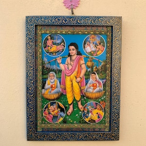 May include: A framed religious print depicting a Hindu deity in a yellow robe holding a staff and a pot. The deity is surrounded by four smaller scenes of other figures. The frame is blue with gold accents.
