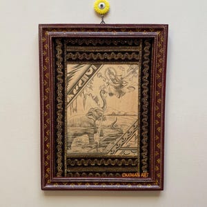 May include: Framed artwork featuring a detailed black and white illustration of an elephant in a river, with a deity above. The frame is a deep red with gold accents and intricate detailing. The artwork is labeled "LAXMAN ART".