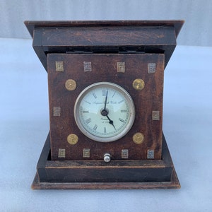 Könnte beinhalten: Eine Holzuhr mit weißem Zifferblatt und schwarzen römischen Ziffern. Das Zifferblatt der Uhr zeigt "Imperial Clock Works" und "Germany Est 1774". Die Uhr ist von kleinen Metallquadraten und -kreisen umgeben.