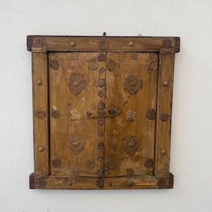 May include: A vintage, rectangular wooden cabinet with two doors. The cabinet is a warm, golden brown color and is adorned with decorative brass accents, including circular and floral designs. The doors are secured with a metal latch. The text "LAXMAN ART" is visible.